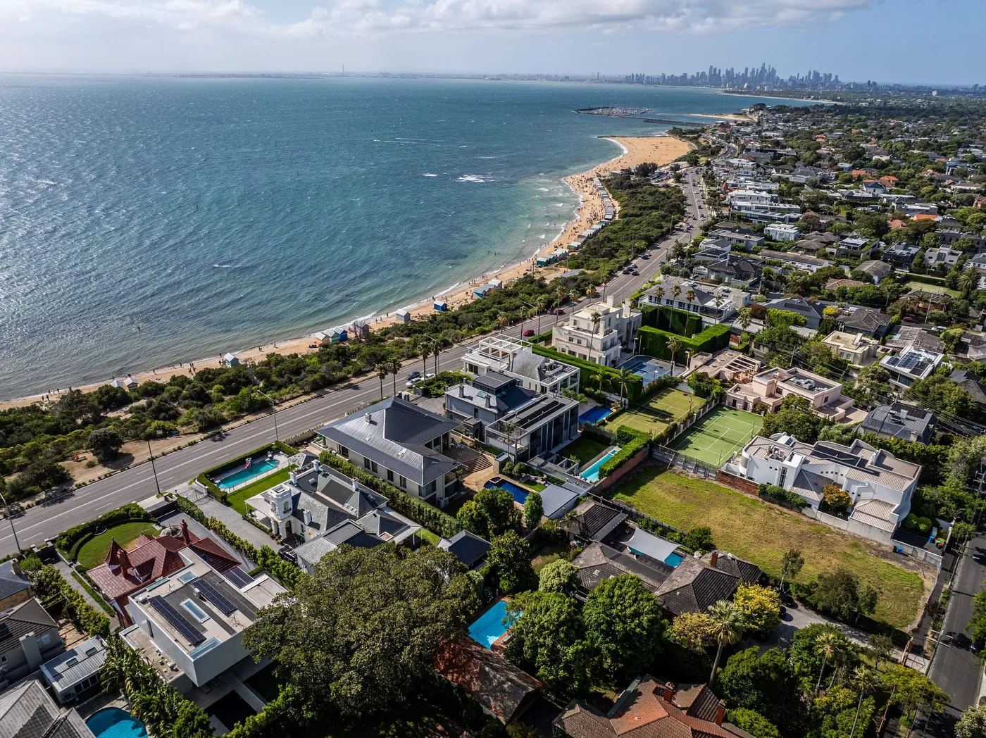 Aerial along a bayside beach and its homes, the Melbourne skyline on the horizon