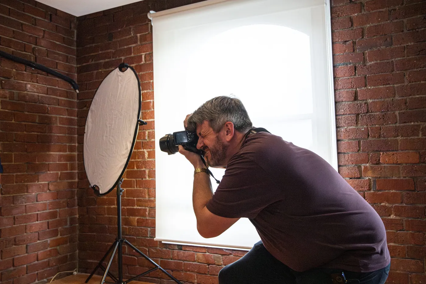 A photographer crouching to shoot beside a reflector in a brick studio